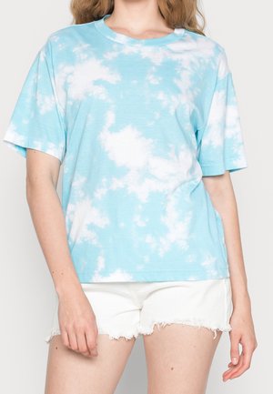 Kobieta ubrana w jasnoniebieską i białą koszulkę z krótkim rękawem w stylu tie-dye oraz białe sprane jeansowe szorty.