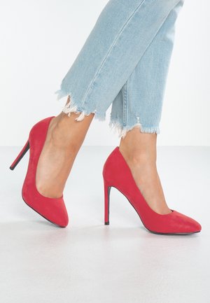 Rote Wildleder-Pumps mit hohen Absätzen, spitz zulaufender Zehenpartie und schlanker Silhouette, kombiniert mit blauen Jeans mit fransigem Saum.