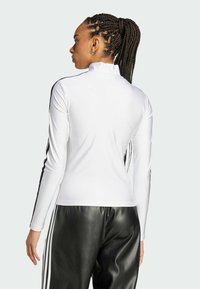 Valkoinen pitkähihainen paita, jossa on mock neck -kaulus ja mustat sivuraidat. Yhdistetty mustiin nahkahousuihin, mikä korostaa sileää pinnan rakennetta ja istuvaa muotoilua.