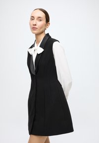 Black sleeveless vest with a fitted design, featuring a notched collar and button closure, paired with a white blouse with a bow tie.
