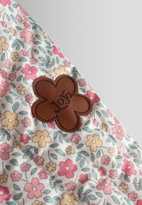 Bruine leren bloemvormige patch met "JoJo maman bébé" op witte stof met een roze, gele en groene bloemenpatroon.
