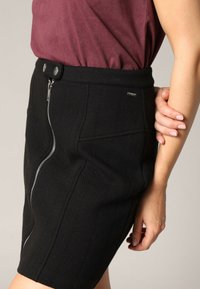 Jupe noire texturée à coupe slim, avec une fermeture éclair sur le côté, deux boutons de fermeture et de subtils détails de couture.