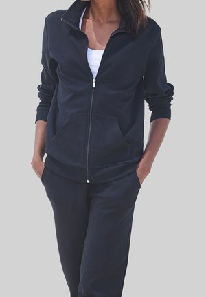 Femme portant une veste noire zippée avec des poches et un pantalon noir assorti, debout, les mains dans les poches, devant un fond uni.