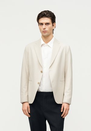 Ung mann med beige blazer over hvit skjorte og svarte bukser, stående mot en ensfarget lys bakgrunn.