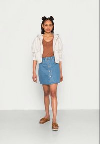 Sweatshirt branco com fecho éclair por cima de uma blusa castanha, combinado com uma saia de ganga azul com botões frontais e sandálias de plataforma castanhas com uma base estampada.