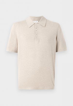 Mladý muž s kučeravými tmavými vlasmi, oblečený v béžové krátkorukávovej polokošeli, hnedých kockovaných nohaviciach, čiernych topánkach, stojí s rukami v vreckách na bielom pozadí.