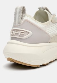 Baskets texturées beige et blanches avec un talon renforcé, une semelle extérieure en caoutchouc et un marquage minimal. Comprend un col en tissu et une languette de traction.