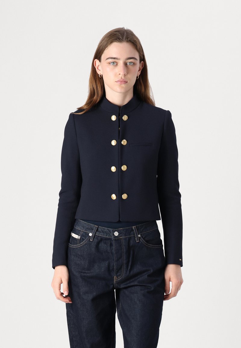Giacca cropped blu realizzata in tessuto liscio, con colletto alla cinese, bottoni dorati e una tasca laterale. Abbinata a pantaloni in denim scuro.
