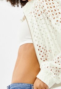 White cropped jacket with open eyelet pattern, zip closure, and gathered cuffs, layered over a white ribbed tank top. Light denim shorts visible.