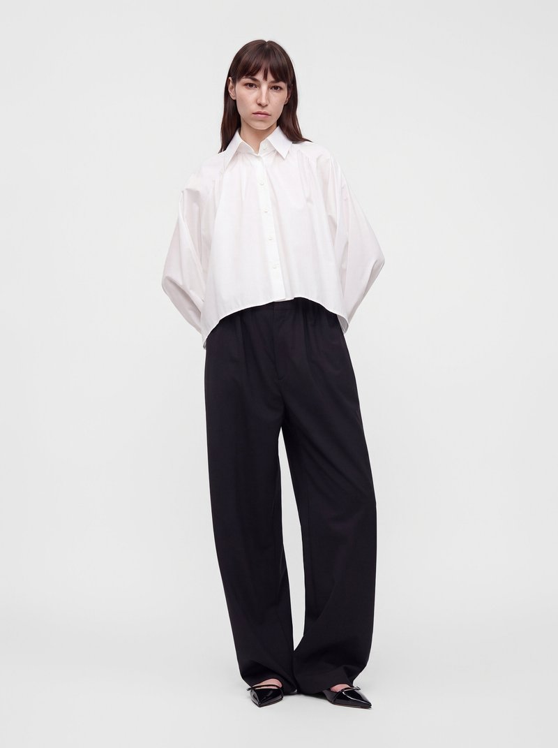 Femme debout portant une chemise blanche ample boutonnée, un pantalon large noir et des chaussures pointues noires sur un fond clair uni.