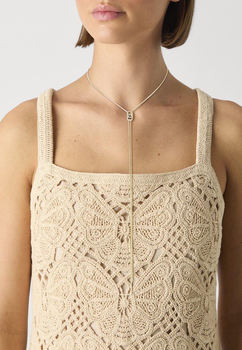Top de punto beige con un patrón de mariposas, con tirantes anchos. Un collar de lariat plateado con un colgante se sostiene en el escote.