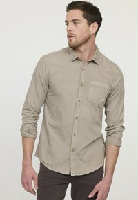 Chemise beige à manches longues avec un boutonnière, dotée d'une poche, présentant une texture douce et un ourlet arrondi. Les boutons sont d'une couleur claire contrastante.
