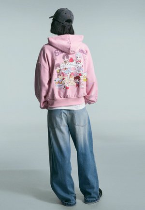 Person wearing a pink hoodie with colorful cartoon characters and a cake on the back, paired with loose blue jeans and a dark cap.