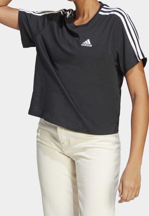 Person trägt ein schwarzes, kurzes Adidas-T-Shirt mit weißen Streifen auf den Schultern und beige hoch taillierte Hose, Arm erhoben.