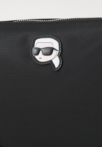 Bolsa de nylon negra con un rostro de caricatura con cabello blanco y gafas de sol como parche en la parte frontal, realzada por un cierre metálico.