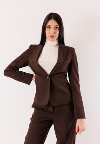 Donna con lunghi capelli scuri che indossa un completo marrone e un dolcevita bianco, in piedi con una mano sul fianco contro uno sfondo semplice.