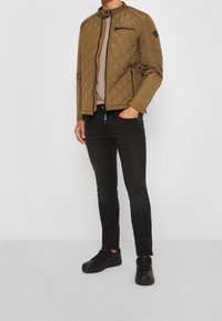 Hnědá prošívaná bunda s vysokým límcem, zipovými kapsami a černými akcenty, spárovaná s černými slim-fit džíny a černými teniskami.