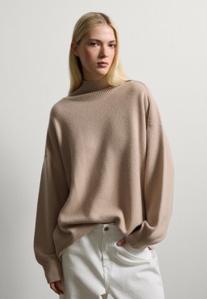 Suéter beige de gran tamaño con un alto cuello acanalado y hombros caídos, combinado con pantalones blancos, presentado sobre un fondo claro.