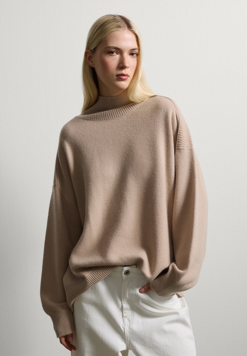 Beige Übergröße-Pullover mit hohem geripptem Kragen und überschnittenen Schultern, kombiniert mit weißen Hosen, vor einem hellen Hintergrund präsentiert.