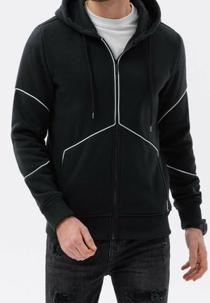 Mężczyzna w czarnej bluzie z zamkiem i białymi lamówkami oraz rozciągniętych czarnych dżinsach, stojący na tle jednolitego tła.