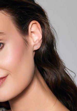 Srebrne kolczyki w kształcie sztyftów z małym, przezroczystym kamieniem szlachetnym. Design jest minimalistyczny, osadzony na tle falujących brązowych włosów.