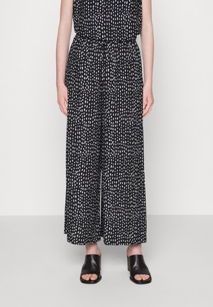 Czarne spodnie o szerokich nogawkach w białe kropki, noszone z czarnymi otwartymi sandałami na obcasie, pokazane od talii do stóp.