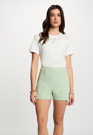 Femme aux longs cheveux noirs portant un haut blanc à manches courtes avec des détails en dentelle et un short taille haute vert clair, debout devant un fond uni.