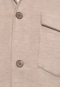 Beige corduroy stof met twee bruine knopen en een klepzak zichtbaar aan de rechterzijde van het kledingstuk.