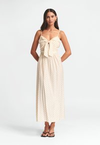 Maxi sukienka w jasnobeżowym materiale z czarnymi kropkami. Posiada detal związywania z przodu, cienkie ramiączka oraz zwiewny krój.