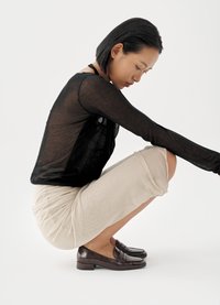 Μαύρο διαφανές τοπ με μακριά μανίκια, μπεζ φούστα μήκους μέχρι το γόνατο και σκούρα καφέ μοκασίνια. Το μοντέλο είναι σε ημικαθιστή θέση με τα χέρια να στηρίζονται στα γόνατα.