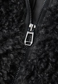 Tessuto in pelliccia sintetica nera con superficie texturizzata e soffice, caratterizzato da una cerniera argento con una targhetta rettangolare e rifinitura in nylon nero.