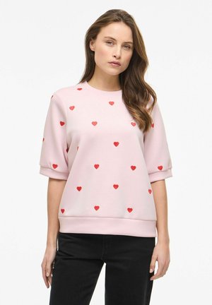 Donna che indossa una maglietta a maniche corte rosa chiaro con motivi a cuore rossi e pantaloni neri, in piedi contro uno sfondo bianco uniforme.