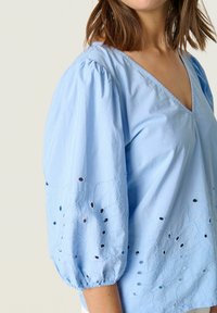 Top in cotone azzurro chiaro con scollo a V, maniche a palloncino e motivi di foglie ricamati. Presenta piccoli tagli e polsini arricciati.