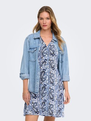 Mujer que lleva un vestido azul con estampado paisley y una chaqueta vaquera clara, de pie con los brazos relajados, expresión facial neutra, fondo claro.