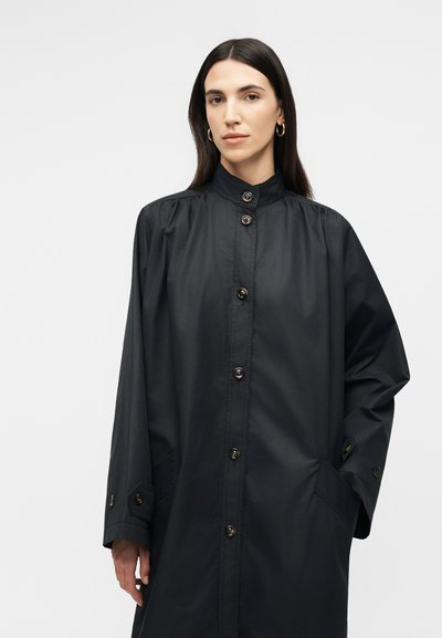 Femme aux longs cheveux foncés portant un manteau noir boutonné avec deux poches devant et un col montant, debout devant un fond clair uni.