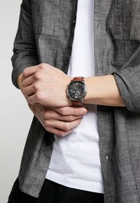 Montre avec un cadran noir, un boîtier en métal argenté, des fonctions chronographe et un bracelet en cuir marron. Portée au poignet, assortie à une chemise grise.