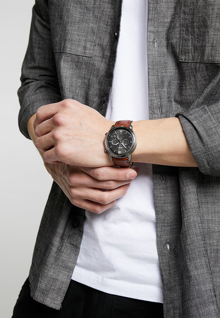Montre avec un cadran noir, un boîtier en métal argenté, des fonctions chronographe et un bracelet en cuir marron. Portée au poignet, assortie à une chemise grise.