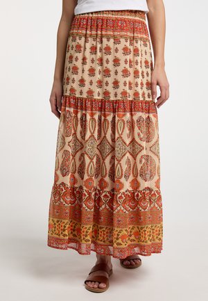 Kvinde iført et langt, fler-mønstret nederdel i røde, beige og gule toner med blomster- og geometriske designs, kombineret med brune sandaler.