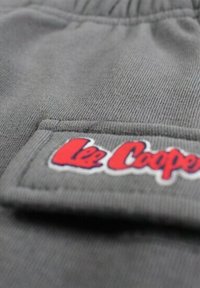 Tissu en coton gris avec une texture visible, comportant un patch blanc brodé de "Lee Cooper" en rouge et bleu. Le design inclut une poche.