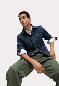Chaqueta de mezclilla en azul oscuro con cuello, bolsillos en el pecho y puños enrollados, combinada con pantalones verde olivo. Textura suave y ajuste entallado.