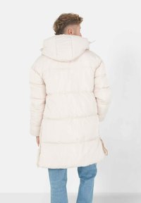 Lichtbeige pufferjack met een capuchon, voorzien van horizontale stiksels, een langere snit en zijsplitten voor meer bewegingsvrijheid.