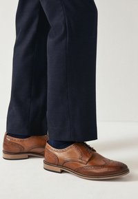 Zapatos brogue de piel marrón con un diseño de cordones, que presentan un patrón perforado y una suela de contraste, combinados con pantalones azul marino oscuro.
