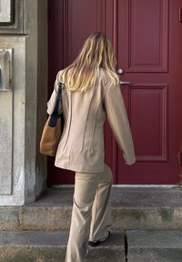 Beige-säätöinen puku, jossa on pystysaumat, yhdistettynä ruskeaan olkalaukkuun ja mustiin kenkiin. Taustalla punainen ovi ja portaat, jotka johtavat ylös.