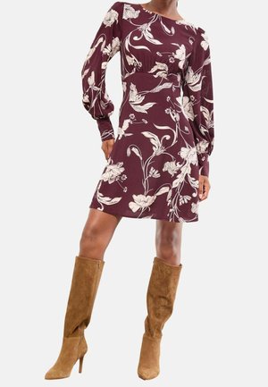 Femme portant une robe fleurie bordeaux à manches longues bouffantes et des bottes à talons marron en suède montant jusqu'aux genoux, debout avec une main sur la hanche.