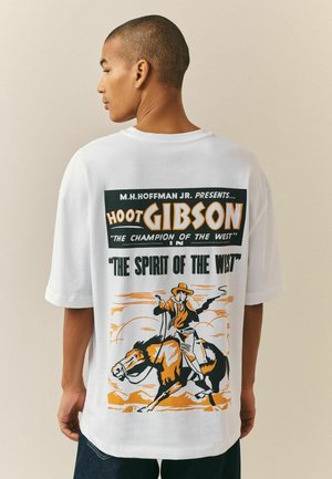 Personne portant un t-shirt blanc avec un graphisme vintage de cowboy orange et noir et un texte sur Hoot Gibson, "Champion de l'Ouest."