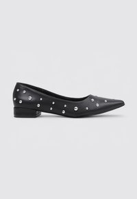 Zapato plano de cuero negro con punta afilada, adornado con tachuelas metálicas y un tacón bajo y cuadrado. Textura suave y diseño elegante.