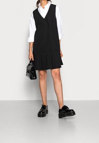 Abito nero senza maniche con gonna plissettata, design doppiopetto, indossato sopra una camicia bianca. Modella che tiene una piccola borsa nera e indossa scarpe con plateau.