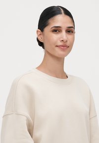Ung kvinne med mørkt hår samlet bakover, iført en lys beige, oversized hettegenser, som står med ansiktet fremover og et svakt smil mot en ensfarget bakgrunn.