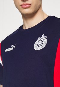 Navy cotton T-shirt featuring red side panels, white Puma logo, and embroidered Club Deportivo Guadalajara crest. Round neck, short sleeves.