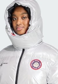 Srebrna pufasta jakna s visokim ovratnikom, koja ima okruglu naljepnicu i crni zatvarač. Tkanina ima sjajnu teksturu i kviltani dizajn.